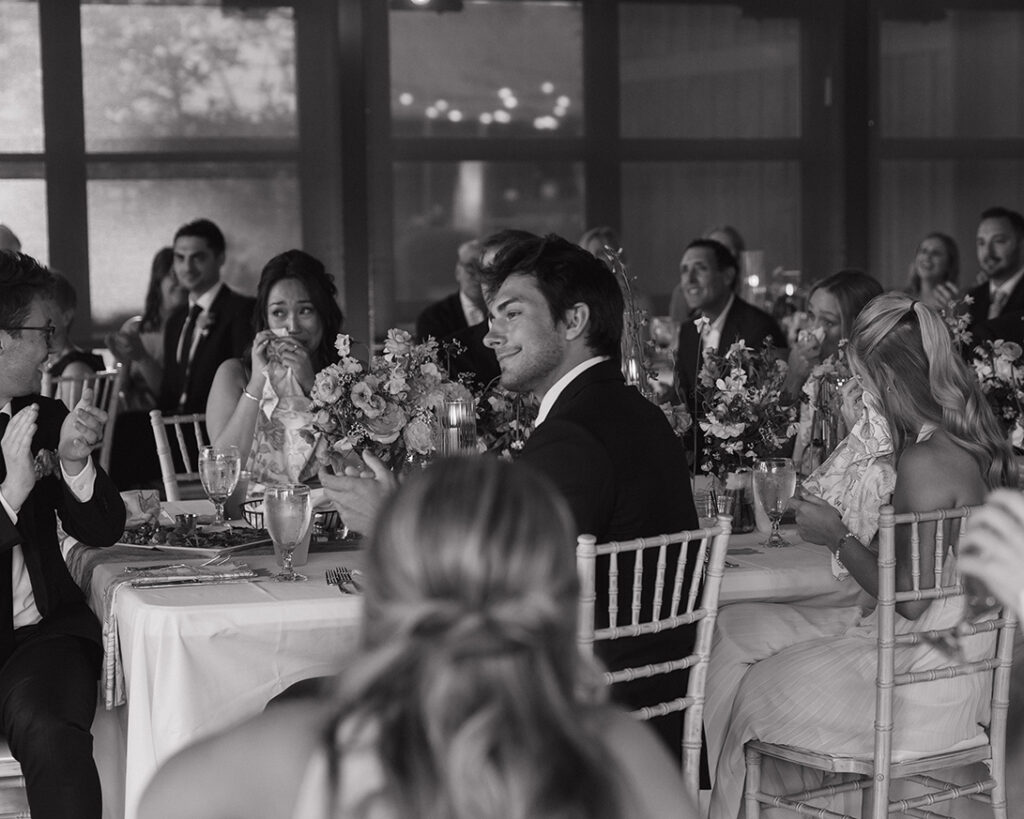 A black-and-white photo of a wedding reception shows guests seated at elegantly decorated tables, smiling and crying from speeches.