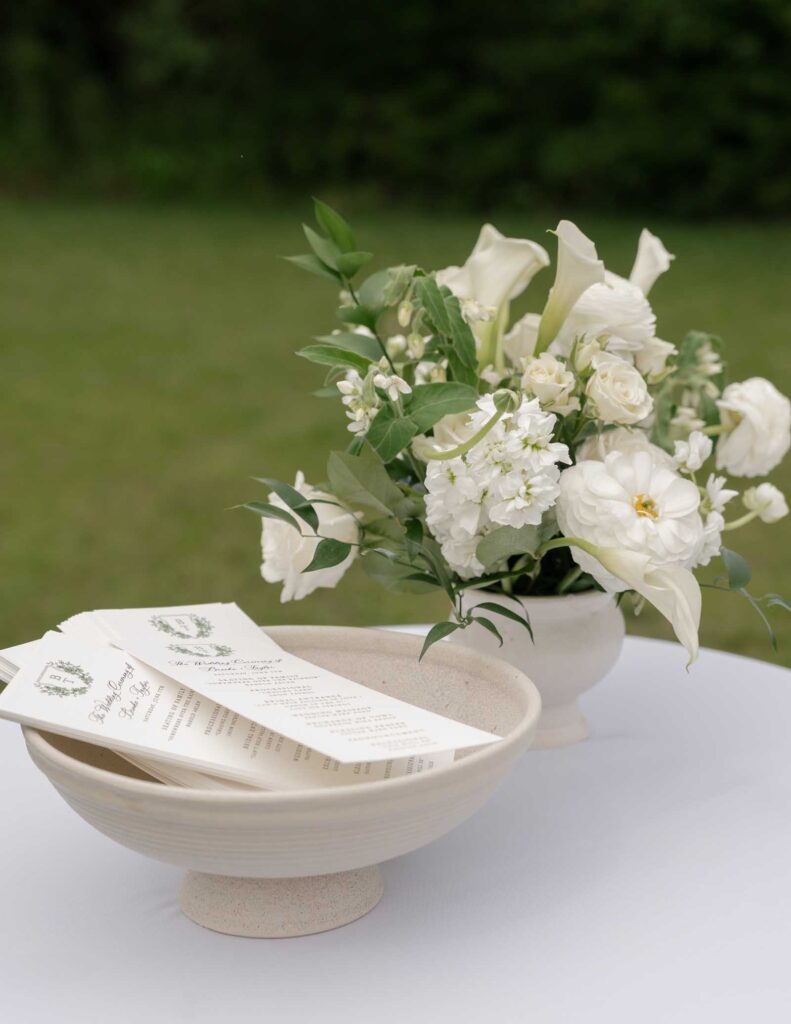 Elegant white calla lily and rose bouquet in a ceramic vase on a table with menus in a matching bowl; lush green garden backdrop.
