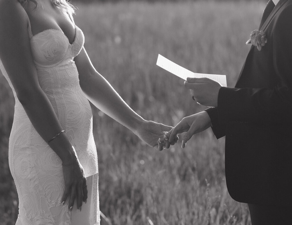 A bride in a lace gown and a suited groom hold hands in a sunlit field, exchanging vows. The scene conveys love and commitment in a serene setting.