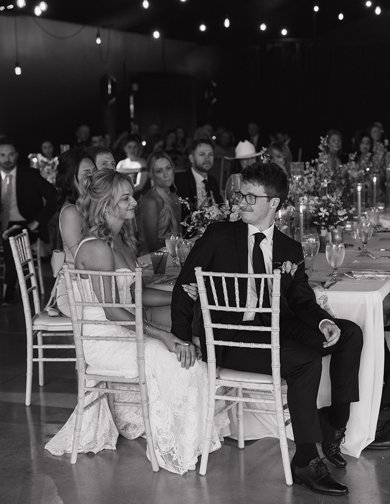 A couple, seated on wooden chairs, shares a loving gaze during a Westerville wedding reception. She's in a lace dress; he's in a suit. Guests and floral table decor surround them.