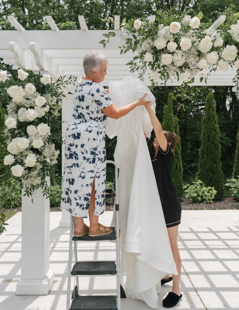 Two women are arranging a white cloth on a pergola decorated with white roses and greenery. One is on a step ladder, and the setting has a serene, elegant atmosphere.