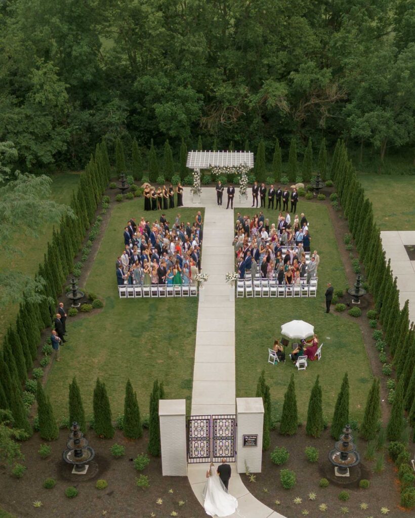 Aerial view of a garden wedding at White Willow Meadows with guests seated on either side of a white aisle. A couple walks toward a floral arch, surrounded by lush greenery.