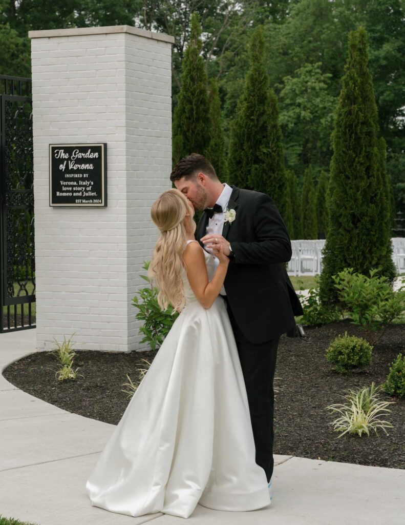 A bride and groom share a kiss outdoors near a white brick column and lush greenery. The bride wears a flowing gown, while the groom dons a black suit.