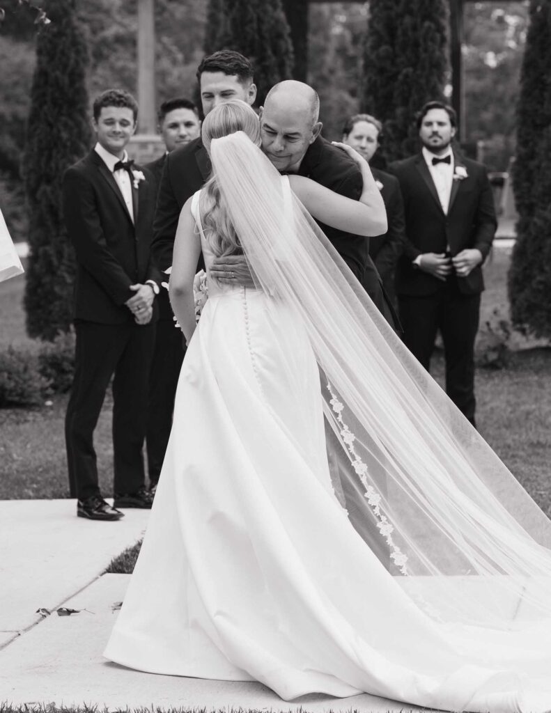 An Ohio bride in a flowing gown and veil embraces an older man in an emotional moment. Groomsmen in suits watch in the background, outdoors in a garden.
