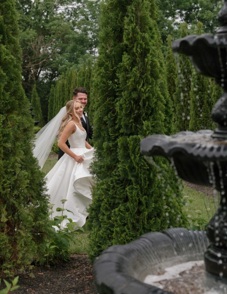 Bride and groom in elegant attire walk through a lush garden at White Willow Meadows, with tall greenery and a nearby stone fountain, conveying joy and romance.