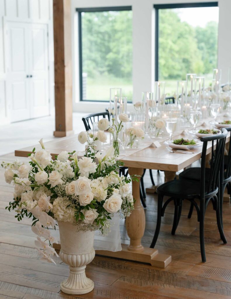 Elegant dining setup with a wooden table adorned with white roses, candles, and black chairs. Large windows reveal lush greenery outside.