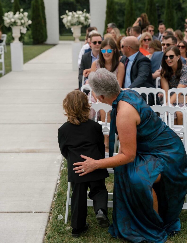 A woman in a teal dress kneels beside a young boy in a suit at an outdoor wedding, looking toward the aisle. Guests sit in white chairs, smiling and chatting.