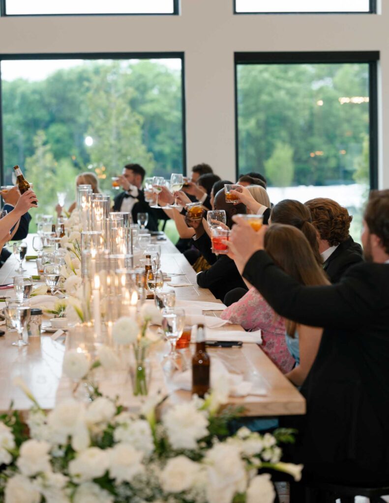 People raise glasses in a celebratory toast at a long table adorned with white flowers and candles. Large windows reveal a scenic green view.