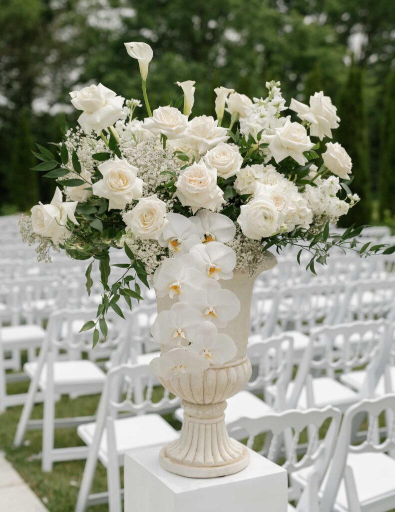 Elegant floral arrangement with white roses, orchids, and greenery in a decorative urn, set in an outdoor venue with white folding chairs at White Willow Meadows in Franklin, Ohio