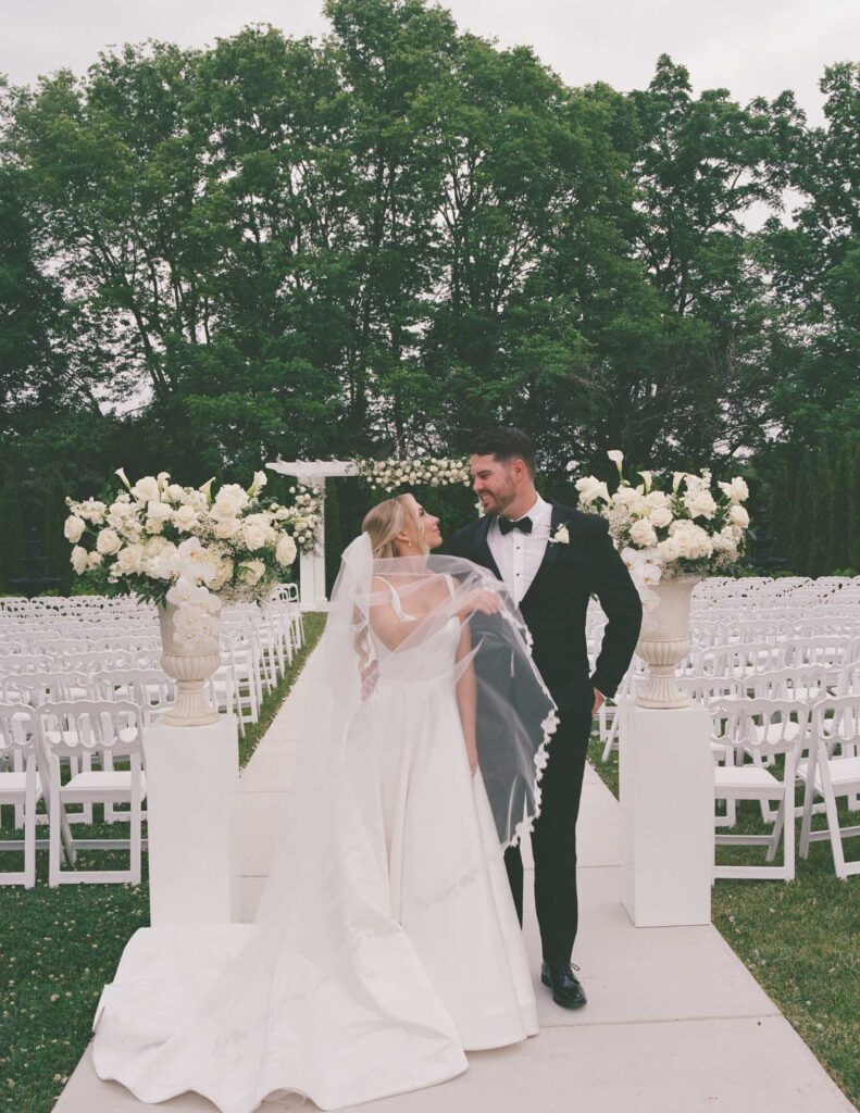 A bride in a flowing white dress and a groom in a black suit smile at each other, walking down an outdoor aisle lined with white chairs and flowers.