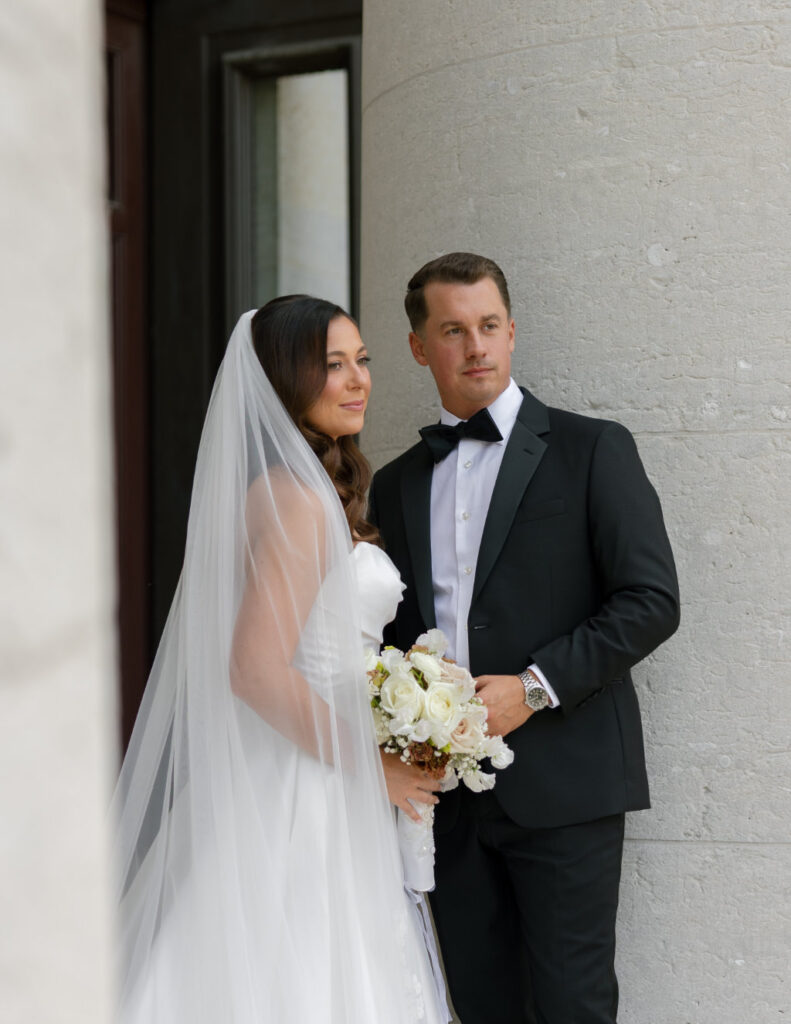 The groom standing confidently in a black tuxedo outside The Ellis with his bride.
