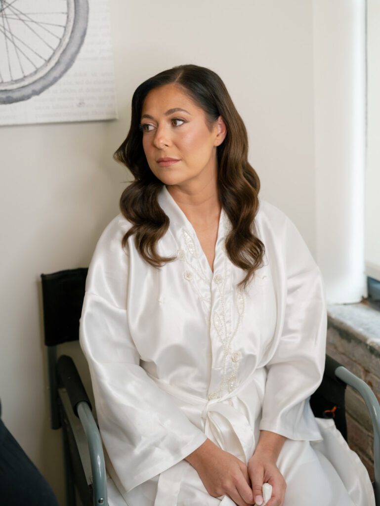 Bride in a white robe soft smiling as makeup artist applies finishing touches.
