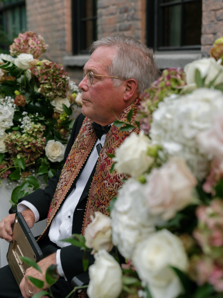 Wedding officiant sitting between floral arrangements.
